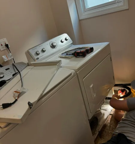 Technician servicing a white dryer beside a washer in a Staten Island laundry room.